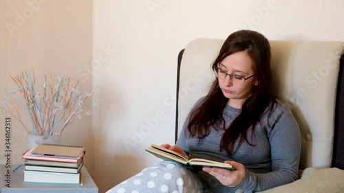 Relaxed woman in eyeglasses and domestic clothing sitting on bed and reading book.  Girl with brown hair relaxing at home with literature.