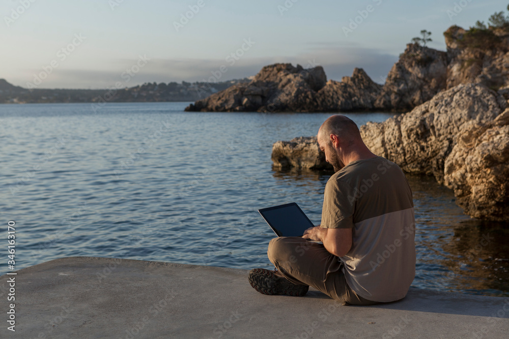 A young man using the laptop at sunrise sitting in the dock on a beach in Mallorca.