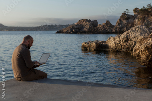 A young man using the laptop at sunrise sitting in the dock on a beach in Mallorca.
