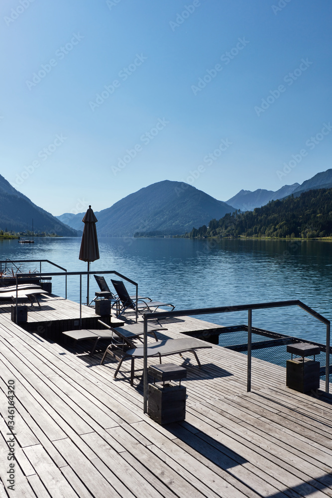 An empty hotel terrace with sun loungers on the shore of a quiet and peaceful alpine lake surrounded by mountains. 