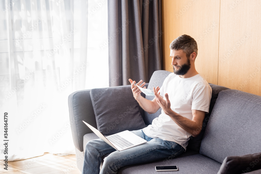 Man holding medical mask near digital devices on sofa in living room Man holding medical mask near digital devices on sofa in living room