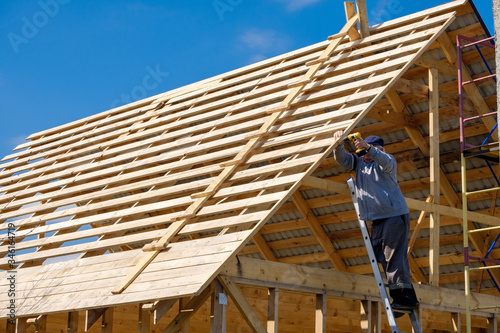 Senior gray-haired builder collects the frame of a wooden country house standing on the stairs against the blue sky. The physical activity of the elderly. Eco-friendly housing made of wood.