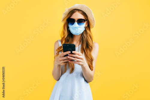 Shocked girl in a summer hat, wears a medical protective mask against a viral infection, uses a mobile phone on a yellow background. Quarantine, coronavirus, summer