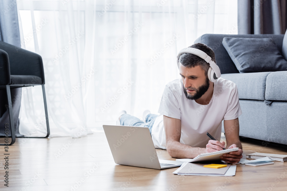 Man in headphones writing on notebook and looking at laptop during webinar in living room Man in headphones writing on notebook and looking at laptop during webinar in living room