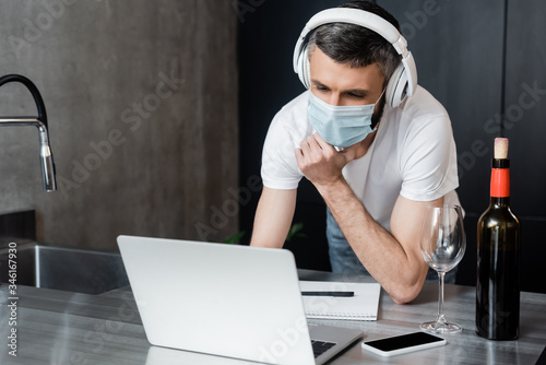 Freelancer in headphones and medical mask looking at laptop near wine and smartphone on kitchen worktop