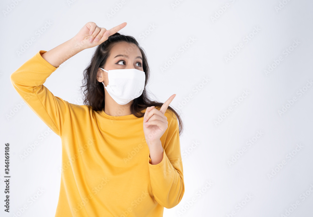 Young happy Asian woman pointing finger hands up to copy space wearing hygienic mask to prevent infection corona virus she wearing yellow sweater shirt shoot in isolated on white background - obrazy, fototapety, plakaty 