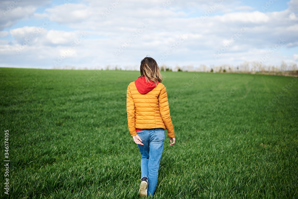 Young blond woman, wearing red hoody, yellow jacket and blue jeans, standing with back to camera on green field in spring. Quarantine getaway in countryside. Stay home, isolate yourself, breathe fresh Young blond woman, wearing red hoody, yellow jacket and blue jeans, standing with back to camera on green field in spring. Quarantine getaway in countryside. Stay home, isolate yourself, breathe fresh