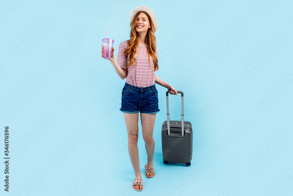 tourist girl in a summer hat standing with a suitcase isolated on a blue background. Travel, summer vacation concept tourist girl in a summer hat standing with a suitcase isolated on a blue background. Travel, summer vacation concept
