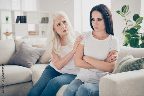 Portrait of two nice attractive lovely pretty charming annoyed irritated disappointed women granny supporting granddaughter sitting on divan in light white interior room house flat apartment