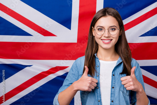 Portrait of positive cheerful foreign student girl enjoy studying abroad courses show thumb up sign wear jeans shirt isolated over british flag background