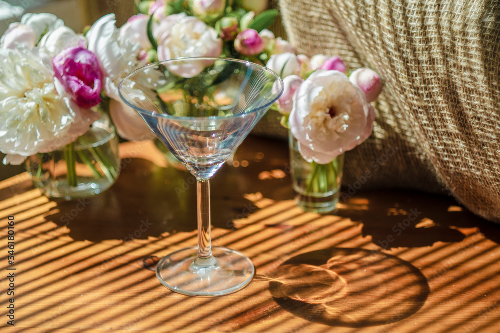 Festive rustic still life with glass empty clear glass surrounded by white flowers on textile background on wooden table. Diagonal striped shadow of the blinds on bright sunny day. Greeting card Festive rustic still life with glass empty clear glass surrounded by white flowers on textile background on wooden table. Diagonal striped shadow of the blinds on bright sunny day. Greeting card