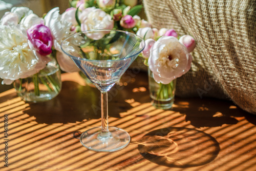 Festive rustic still life with glass empty clear glass surrounded by white flowers on textile background on wooden table. Diagonal striped shadow of the blinds on bright sunny day. Greeting card