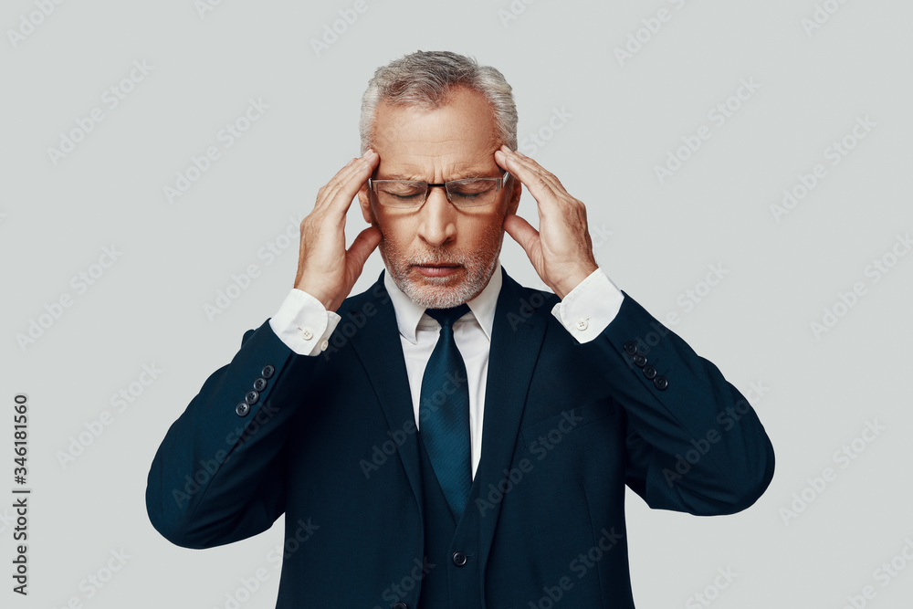 Frustrated senior man in full suit suffering from headache while standing against grey background Frustrated senior man in full suit suffering from headache while standing against grey background