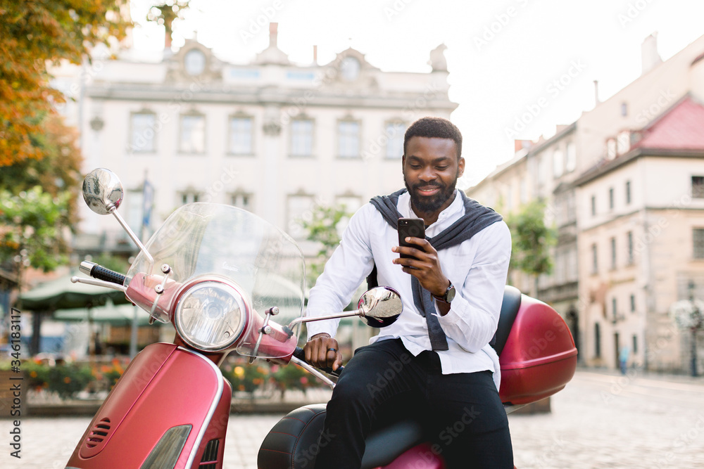 Portrait of smiling young bearded african american guy in smart casual wear  using his smartphone, while sitting on the red scooter outdoors on the old  city square. Urban lifestyle, business foto de
