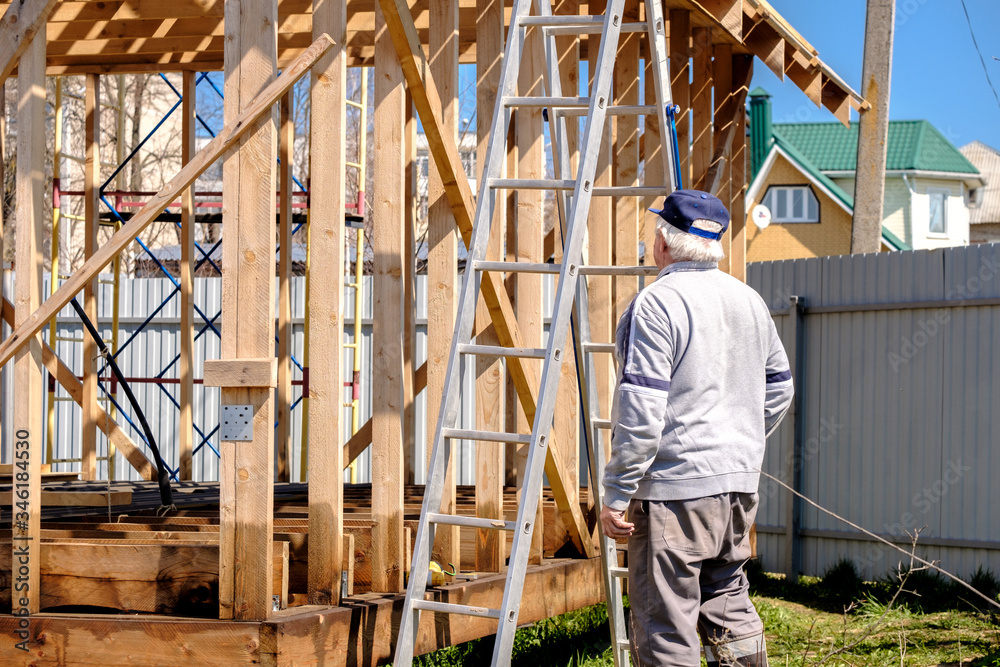 Senior gray-haired builder inspects the frame of a wooden country house under construction against a blue sky. The physical activity of the elderly. Eco-friendly house made of wood. Senior gray-haired builder inspects the frame of a wooden country house under construction against a blue sky. The physical activity of the elderly. Eco-friendly house made of wood.