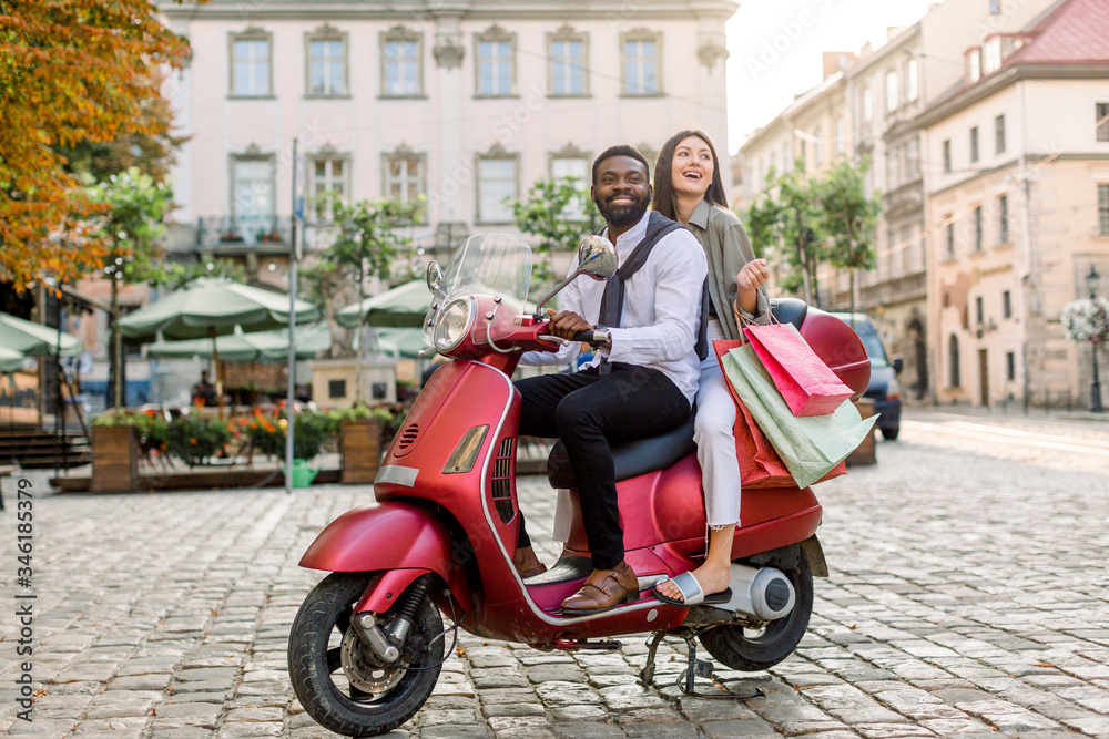 Young beautiful multiracial couple, African guy and Caucasian girl, riding on red motorbike on the city street, happy woman holds shopping bags. Summer Europe vacation, traveling concept