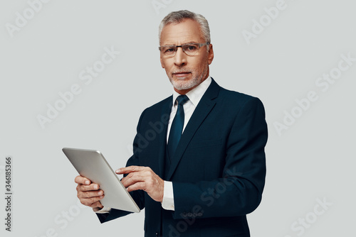 Handsome senior man in full suit using digital tablet and looking a camera while standing against grey background