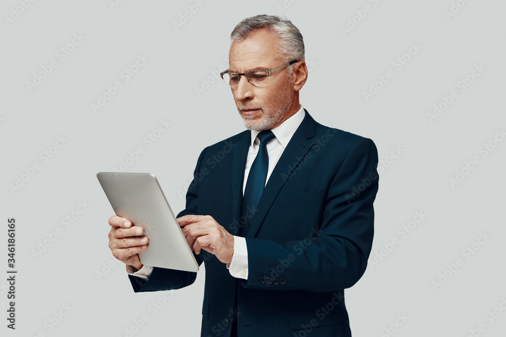Handsome senior man in full suit using digital tablet while standing against grey background Handsome senior man in full suit using digital tablet while standing against grey background