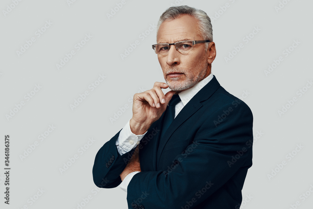 Handsome senior man in full suit looking at camera and keeping hand on chin while standing against grey background Handsome senior man in full suit looking at camera and keeping hand on chin while standing against grey background