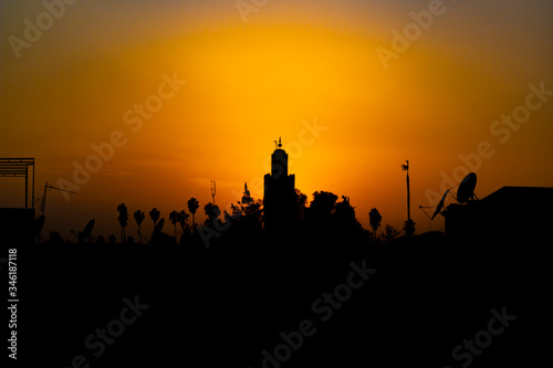 Orange sunset above the minaret of the mosque of Kutubiyya, Marrakesh. Black silhouettes by sunset showing minaret, numerous television antennas and palm tree. Muslim evening prayer time