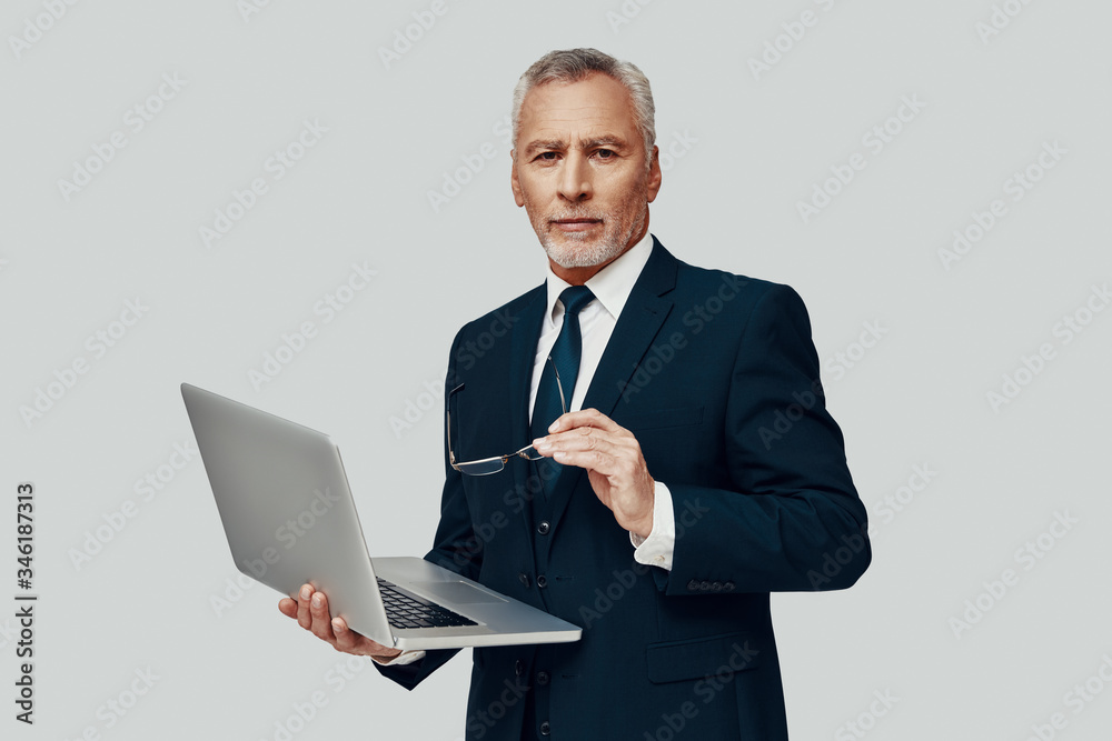 Handsome senior man in full suit using laptop and looking at camera while standing against grey background Handsome senior man in full suit using laptop and looking at camera while standing against grey background