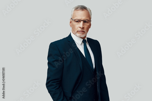 Handsome senior man in full suit looking away while standing against grey background