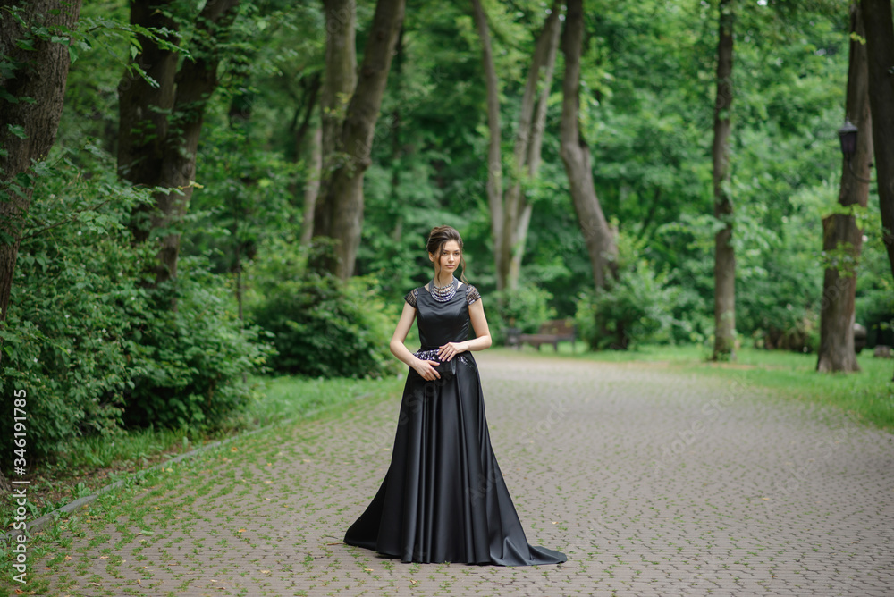 Young beautiful woman posing in a black dress in a park. Holding a handbag in his hands. Young beautiful woman posing in a black dress in a park. Holding a handbag in his hands.