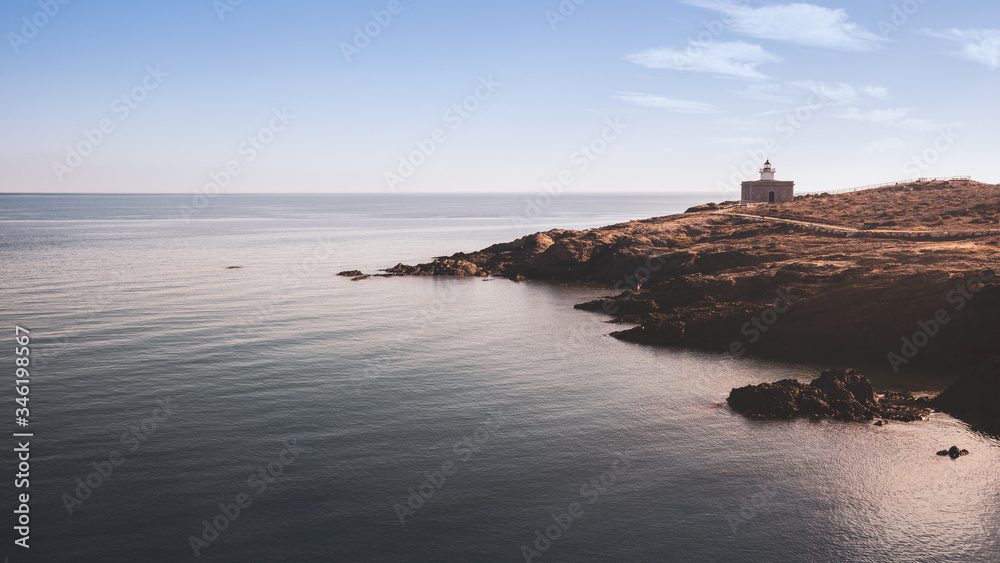 Lighthouse on the rocks of quiet shore of the sea with calm water and blue clear sky. Horizontal view.