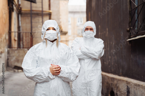 Doctors in protective suits and protective masks stand on the street with serious faces. Walking along the street, guy leads woman foward - they are very determined. The concept of coronirus covid-19 photo