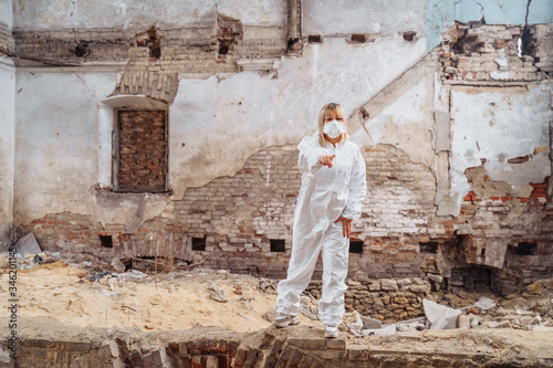 Female medic in a protective suit and medical mask standing on the ruins of a house that was bombed. She pointing finger at the camera and looking forward. She is determined