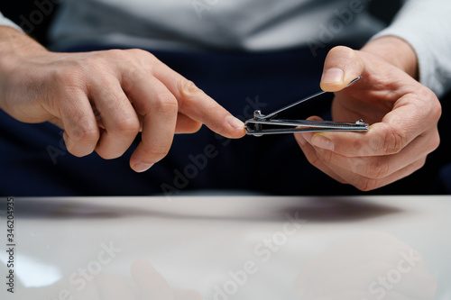 Close up on midsection on hands of unknown caucasian man male holding metal nail clipper clipping cutting nails ba the white table at home trimming and shaping manicure front view