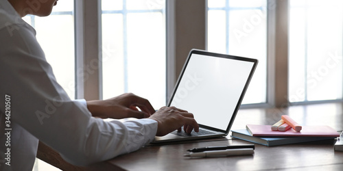 Photo of smart man working as accountant while sitting and working with white blank screen computer laptop at the wooden working desk that surrounded by office equipment over windows as background.
