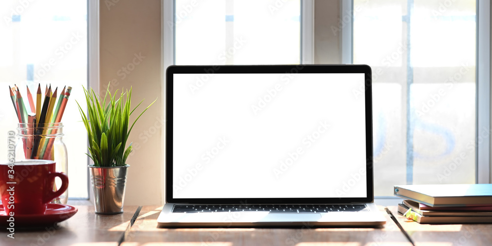 Computer laptop with white blank screen putting on wooden working desk that surrounded by coffee cup, stack of books, potted plant, pencils over comfortable living room windows as background. Computer laptop with white blank screen putting on wooden working desk that surrounded by coffee cup, stack of books, potted plant, pencils over comfortable living room windows as background.