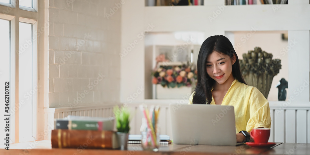 Photo of attractive woman staring on computer laptop that putting on wooden working desk and surrounded by potted plant, coffee cup, stack of books and pencils in vase over sitting room bookshelf.