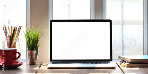 Computer laptop with white blank screen putting on wooden working desk that surrounded by coffee cup, stack of books, potted plant, pencils over comfortable living room windows as background.