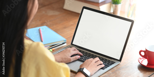 Behind shot of beautiful woman working as accountant while sitting and working with white blank screen computer laptop at the wooden working desk that surrounded by coffee cup and stack of books.