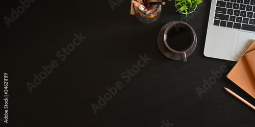 Office desk laptop with black empty screen putting on black working desk that surrounded by coffee cup, potted plant, pencil, notebook and stationary in glass vase.