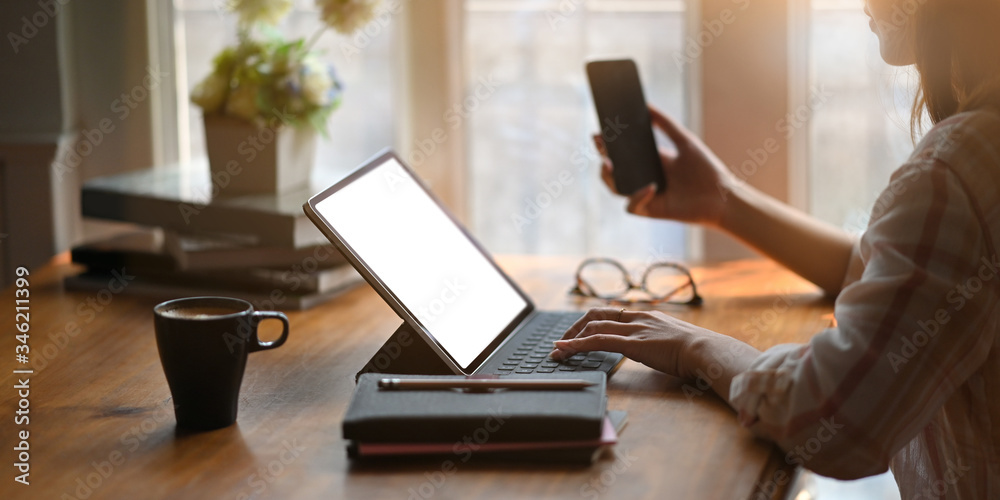 Cropped image of beautiful woman working with white blank screen computer tablet and holding a smartphone in hand while sitting at the wooden working desk over comfortable living room as background. Cropped image of beautiful woman working with white blank screen computer tablet and holding a smartphone in hand while sitting at the wooden working desk over comfortable living room as background.