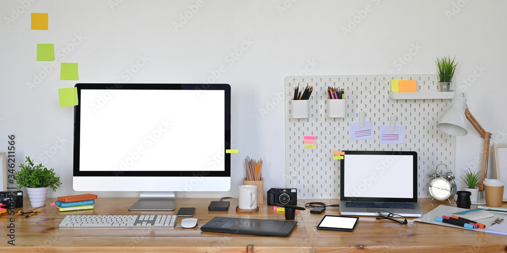 Photo of computer laptop and computer monitor with white blank screen putting on wooden working desk that surrounded by graphic designer equipment over white living room wall as background. Photo of computer laptop and computer monitor with white blank screen putting on wooden working desk that surrounded by graphic designer equipment over white living room wall as background.
