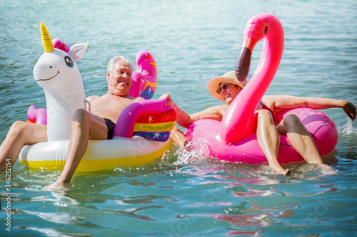 Elderly couple having fun on inflatable flamingo and unicorn. Funny active pensioners happy together enjoying summer vacation on the beach in Europe, laughing, playing the fool, splashing water.