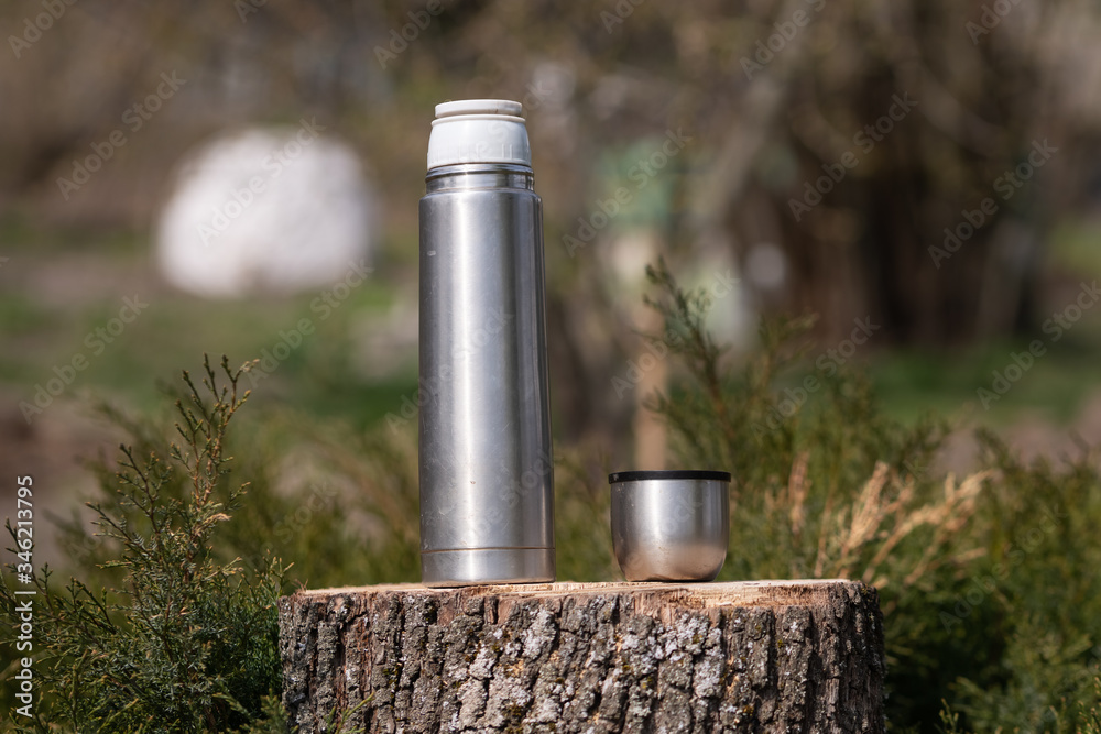 Steel thermos with a cup of hot tee standing on old stump in coniferous forest, camping close up abstract background