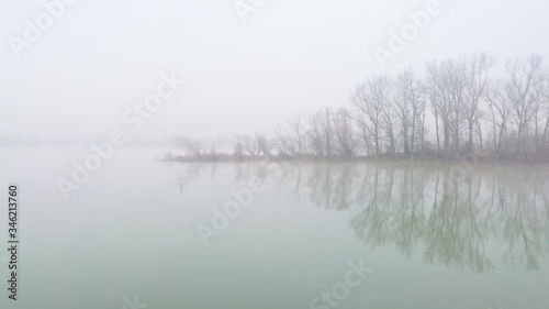 River island in dense fog, with tree silhouettes of bare branches in late fall. The usual autumn morning by the river. Mystical and melancholic autumn landscape.