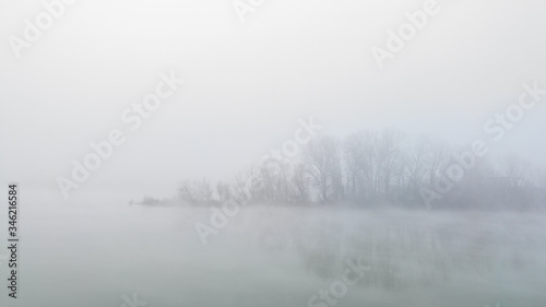 Peaceful autumn scene of mystical river island with a deciduous grove in deep fog early in morning. Calm and silent nature scene covered with thick fog.