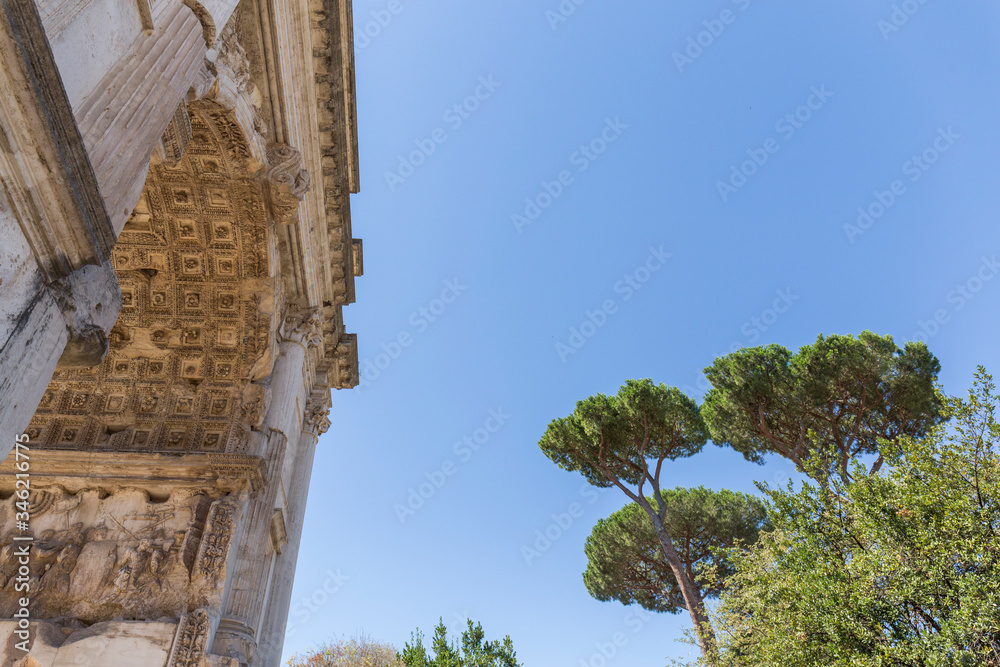 part of a Roman building made of stone, arch seen from below with different engravings in the stone, next to it there is a group of trees that gives color to the photo, the sky is blue