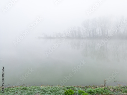 The gloomy landscape of the morning mist in the river valley. A thick fog hovers over the flat surface of the river and creeps among the trees in the woods on a barely visible river island.