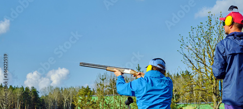 trap shooting - athlete in a wheelchair shoots clay pigeons under the guidance of a shooting trainer, spring blue sky and green forest in the background, Images with Copy Space