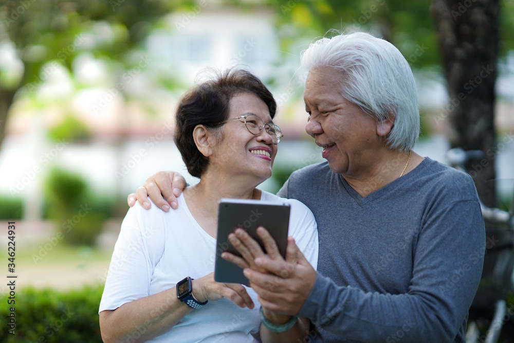 Portrait of happy Asian elderly couple holding tablet together, senior couple using tablet in the park/ yard 