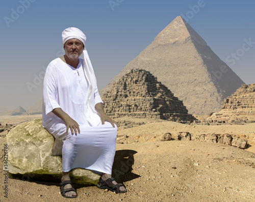 An ancient Egyptian sits on a rock in front of the historic Giza pyramids in Egypt. Right behind him are two little crumbled pyramids. The man is wearing a white robe and a turban.