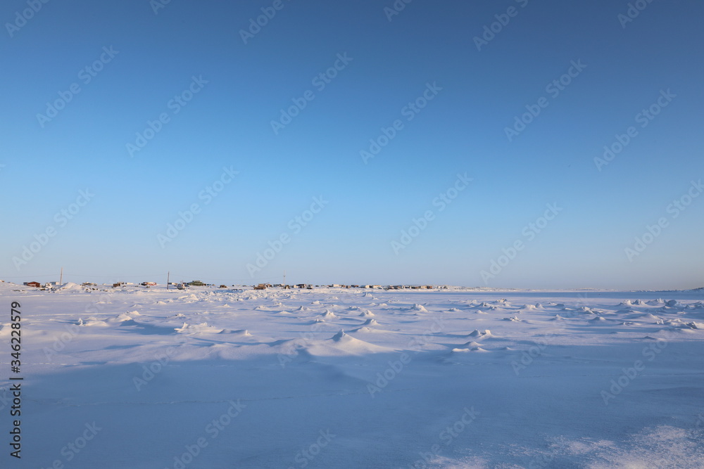 Winter view of an isolated arctic community with snow and ice in the foreground and blue skies in the background, in Arviat, Nunavut Canada