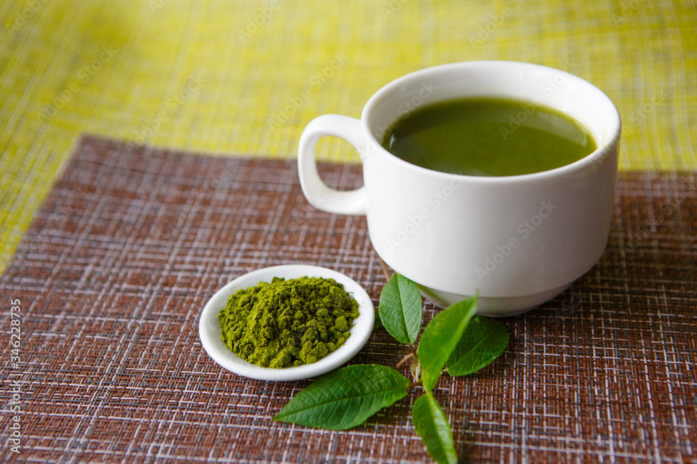 Japanese matcha green tea is poured into a white mug and on a white saucer in powder. Tea set on a textured napkin of natural flowers, decorated with a branch of green leaves. Background with space fo Japanese matcha green tea is poured into a white mug and on a white saucer in powder. Tea set on a textured napkin of natural flowers, decorated with a branch of green leaves. Background with space fo